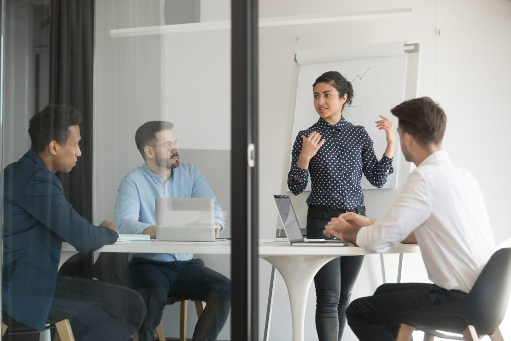 Board room view through glass door diverse businessmen listens hindu firm representative give presentation make offer to investors. Staff takes part in seminar, corporate trainer teach workers concept