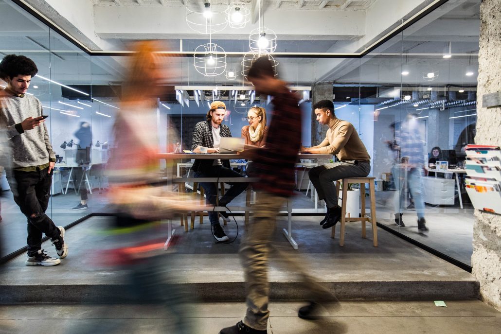 Group of young freelancers working on wireless technology at casual office. Some people are in blurred motion.