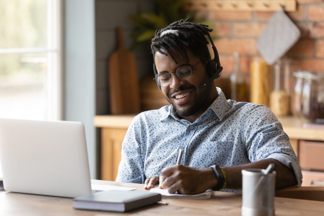 Smiling young African American man in headphones glasses make note watching webinar on laptop. Happy biracial male in earphones work take online course distant training on computer at home.