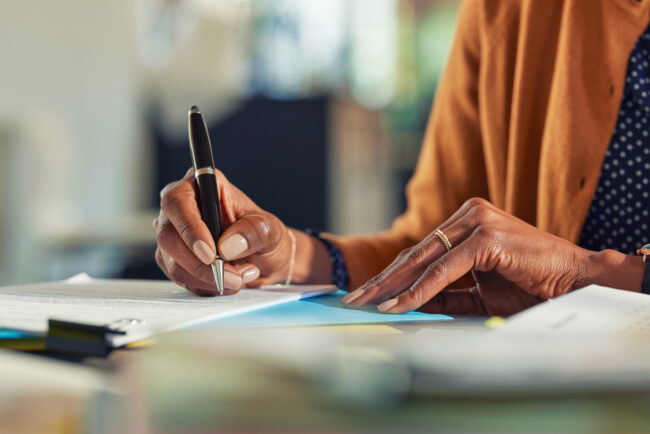 Close up of african american business woman hands writing on report while working in office. Successful black woman signing documents at home using a pen. Mid adult freelancer signing contract document on table.