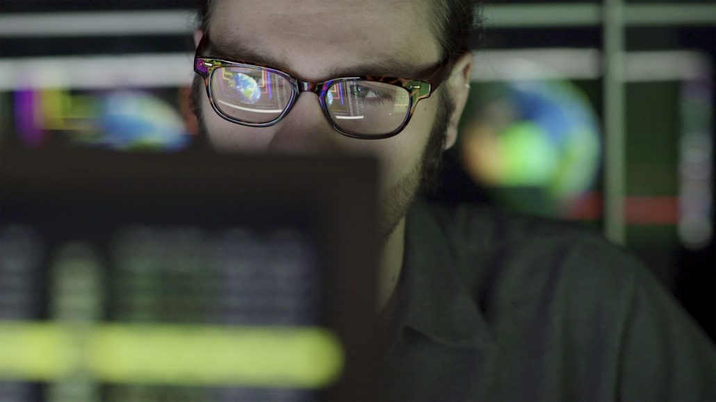 A bearded young man studying a computer screen displaying images of the earth with moving graphics. He’s surrounded by out of focus, colourful displays and reflections showing moving global graphics that are out of focus.