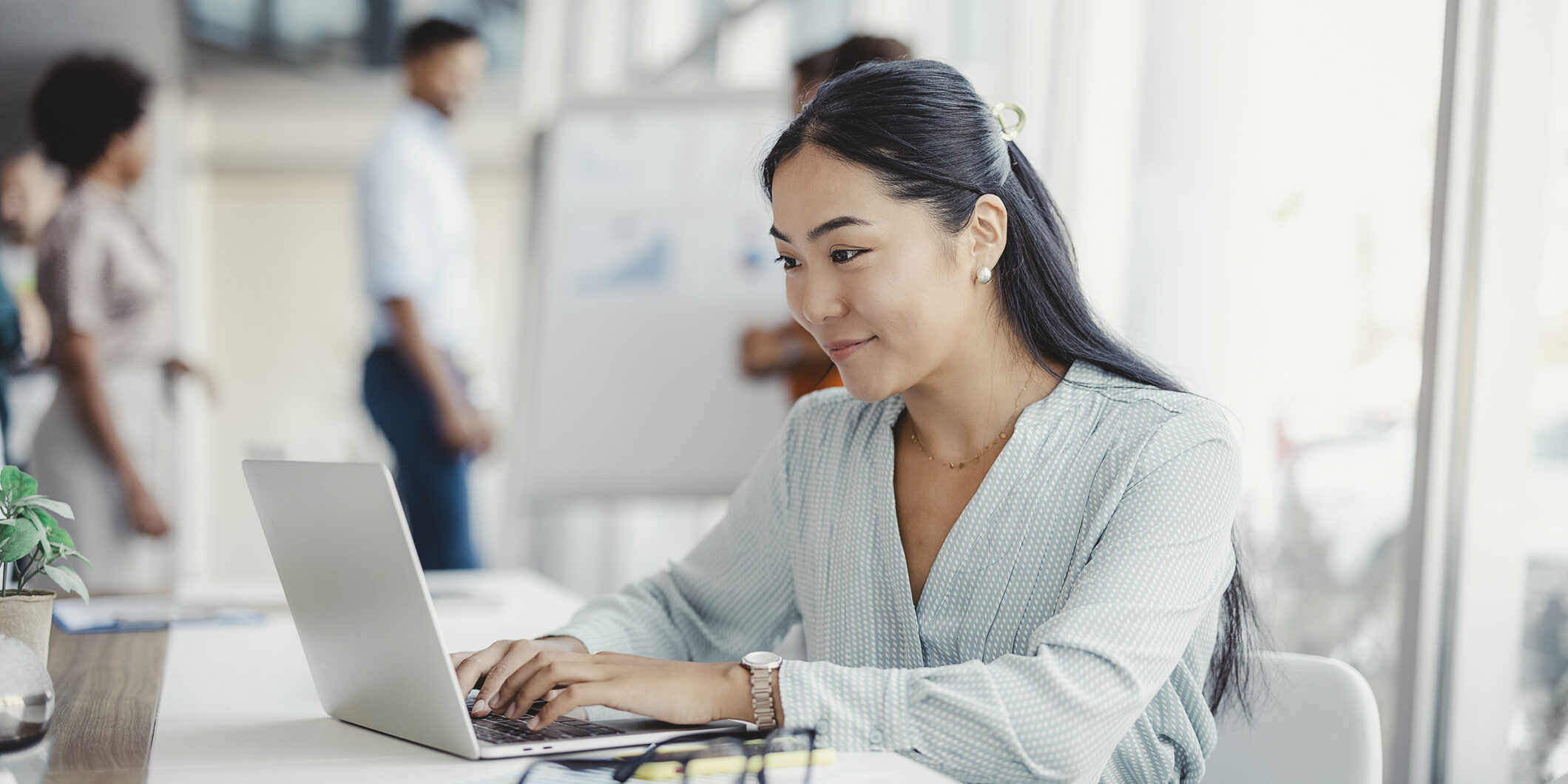 Asian woman working on laptop. Businesswoman busy working on laptop computer at office with colleagues in the background.
